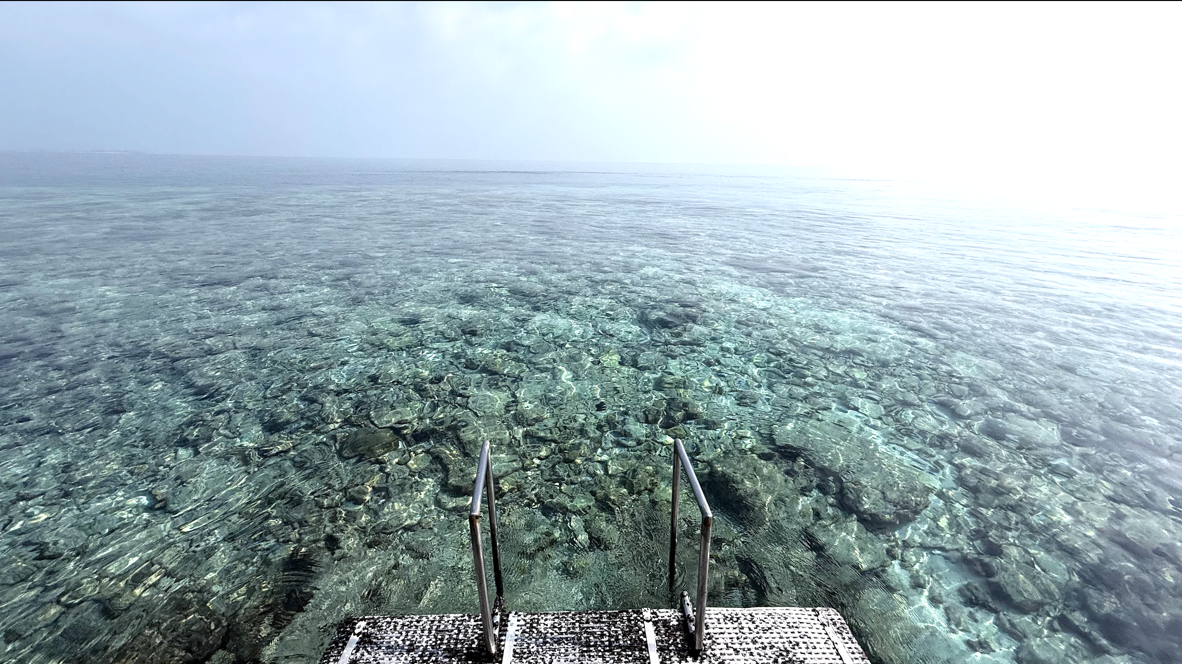 A serene view of clear water and coral formations from a staircase leading into the ocean.